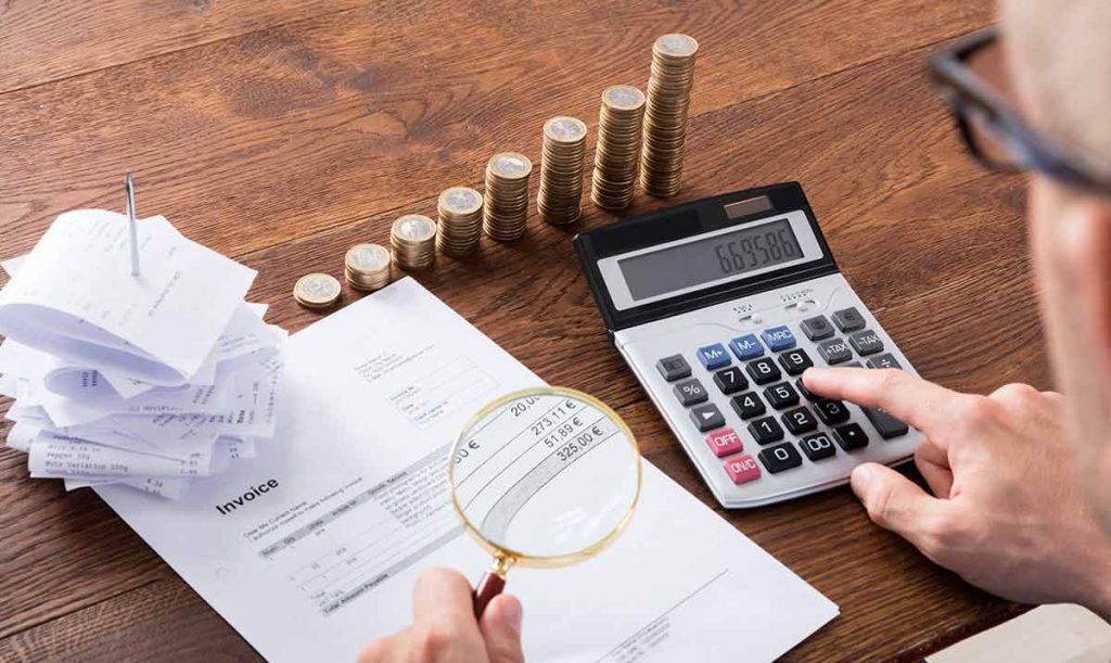 Primo piano della mano di un imprenditore che analizza la fattura con lente d'ingrandimento mentre, con l'altra mano, digita effettua il calcolo delle spese della fattura elettronica con una calcolatrice