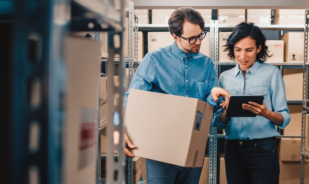 Donna mostra le informazioni su un tablet ad un uomo che tiene una scatola di cartone, sullo sfondo un magazzino di pacchi pronti per la spedizione.