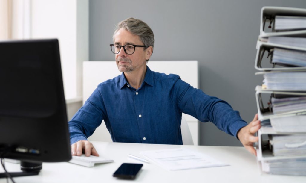 Uomo davanti al pc che spinge via i documenti cartacei e che lavora con i documenti digitali sul computer