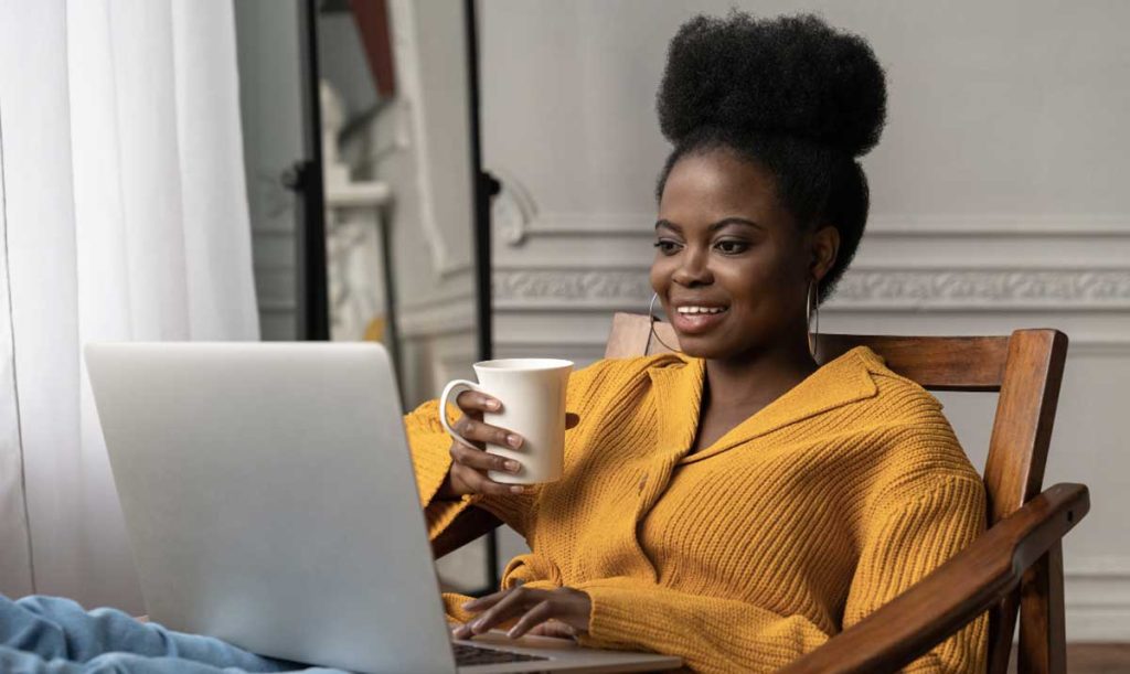 Felice donna afro-americana con acconciatura afro e cardigan giallo, seduta su una sedia, consulta il suo computer portatile mentre beve un tè o caffè.